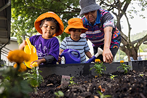 Two young students and a teacher standing in front of a garden bed watering yellow flowers